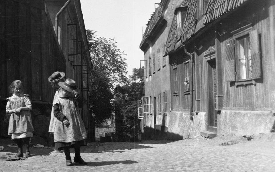 Flickor i gammeldags kläder står på en kullerstensgata bland trähus.